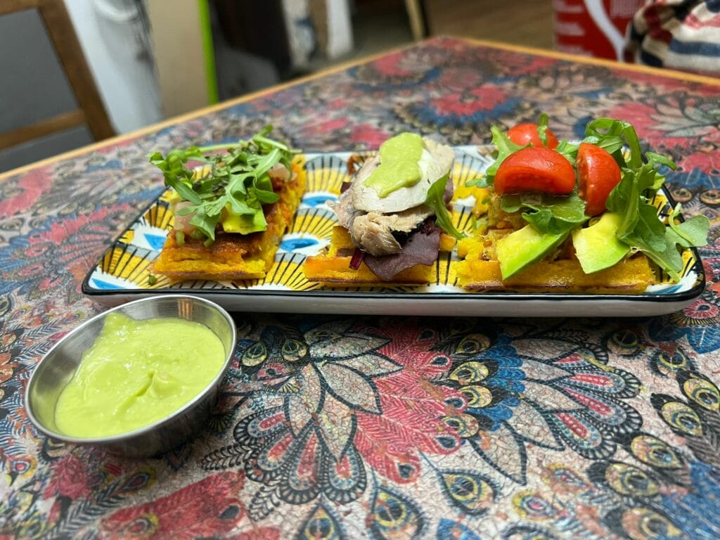 Plate of three types of tapas - one has avocado and tomato, one has slices of fish or meat and the other is topped with greens. A small bowl of sauce is on the side.