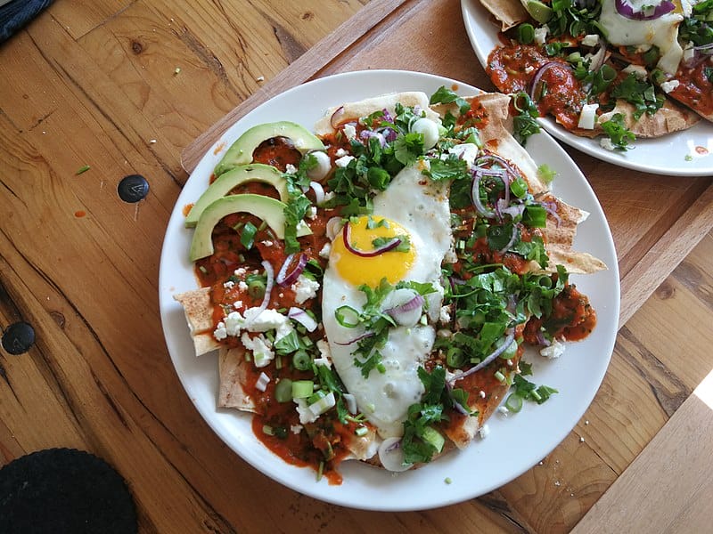 Plate of chilaquiles with red sauce, avocado slices, fried egg topped with queso fresco, red onion and lots of cilantro.