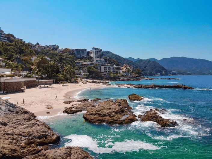 Small sandy beach framed by large rocks and waves, with beachfront buildings in background