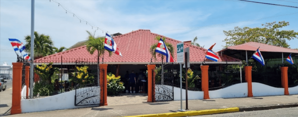 Outside of restaurant - red clay roof open sides to interior of restaurant. Low white fence surrounding building with 4 flags on poles lining the front. You can see a cruise ship docked in the background