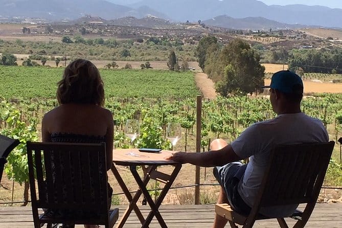 Couple sitting at a little wooden table on a deck looking out over a vineyard with rolling hills in the background.