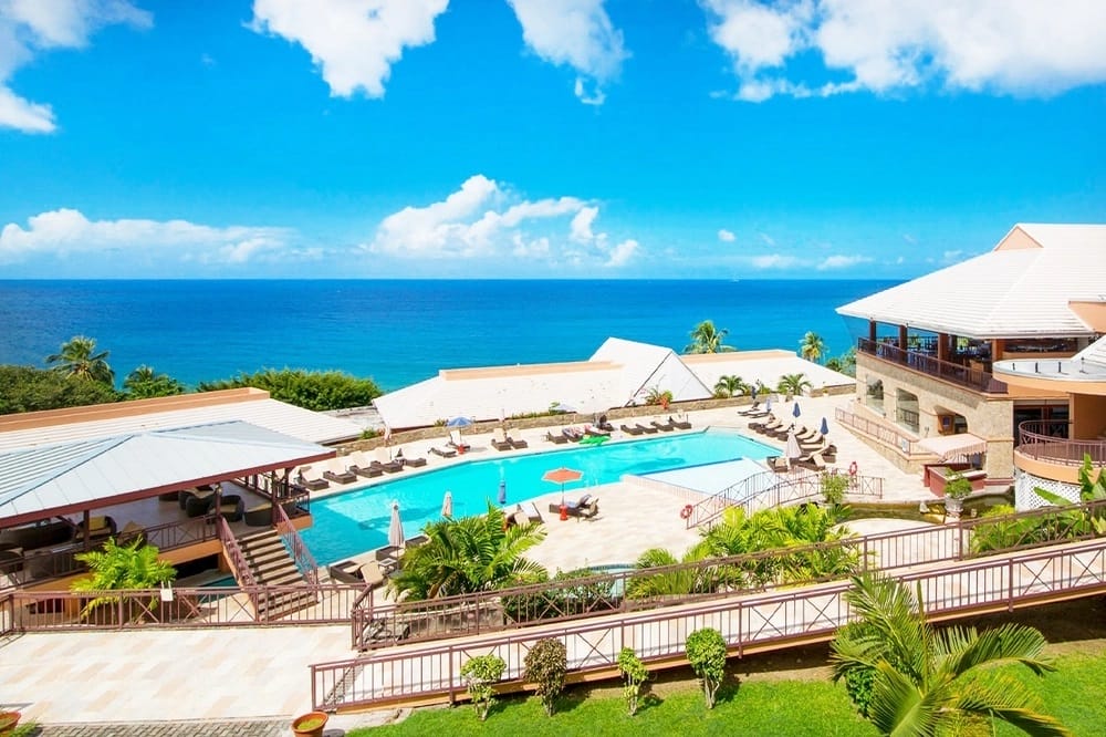 Aerial view of long, rectangular pool surrounded by cabanas and loungers, resort buildings surround the pool. The ocean is in the background.