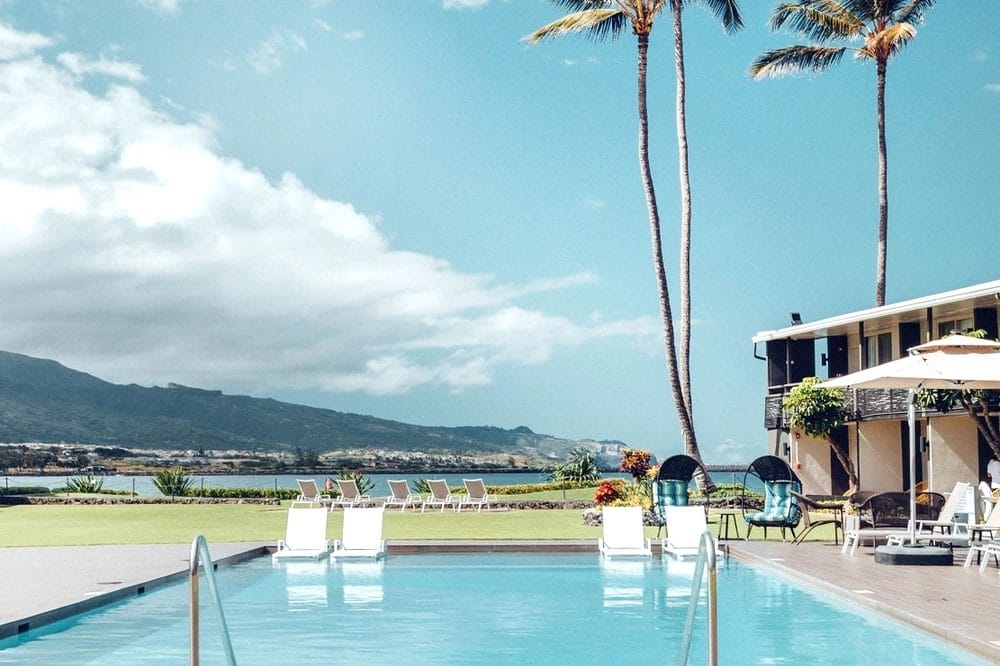 The Maui Seaside Hotel looking from pool, with in-pool loungers out to the bay. Two tall palm trees are beside a two-storey resort building.