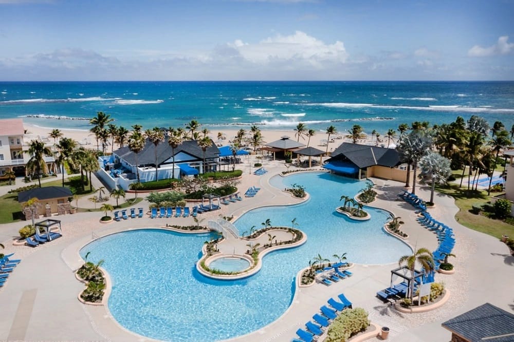 Aerial view of curvy resort pool surrounded by lounge chairs, palm trees and a beach side building. The beach is on the far side of the pool