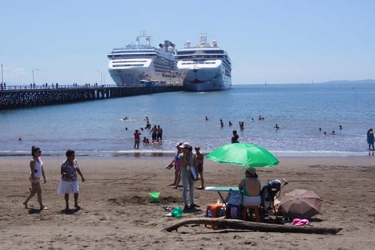 From the shore looking out to sea - in the foreground is the beach with people sitting under an umbrella and a handful of people in the water. To the left of the beach is the cruise ship pier with two ships docked at the end of the pier.