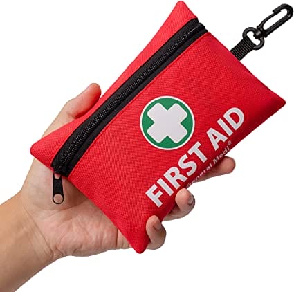 Hand holding a small red bag with a black zipper and black clip to attach to a bag. On it says "First Aid" is block white letters.