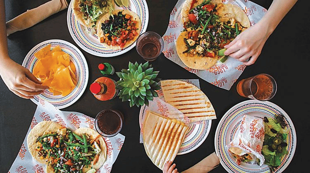Top-down view of table with 4 plates of food - tacos, burritos, quesadillas, 2 bottles of hot sauce and glasses of cola.