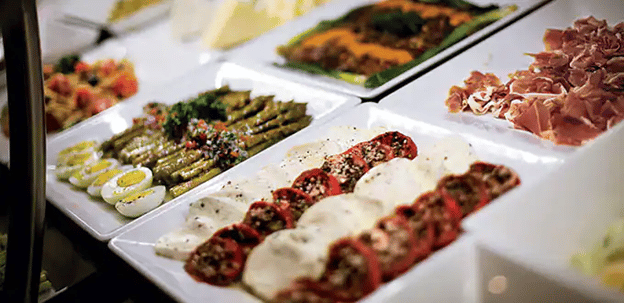 White plates lined up. One has sliced fresh mozzarella and sliced tomatoes drizzled with oil and herbs, another has asparagus spears and hard-boiled eggs cut in half, another has thinly sliced cured meat.
