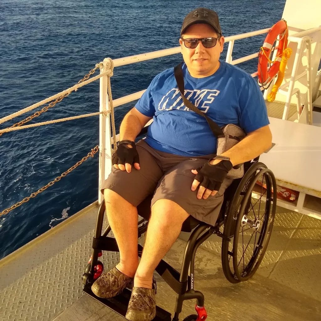 A man sitting in a wheelchair on a cruise ship near the railing. He is wearing a blue t-shirt and a ball cap and sunglasses. He is a nice man.