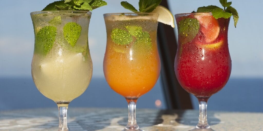Three colorful drinks in stemmed glasses and fruit and garnishes in the glass and the water in the background.