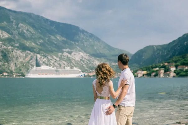 A couple standing on the shore looking out toward a cruise ship docked across the bay.
