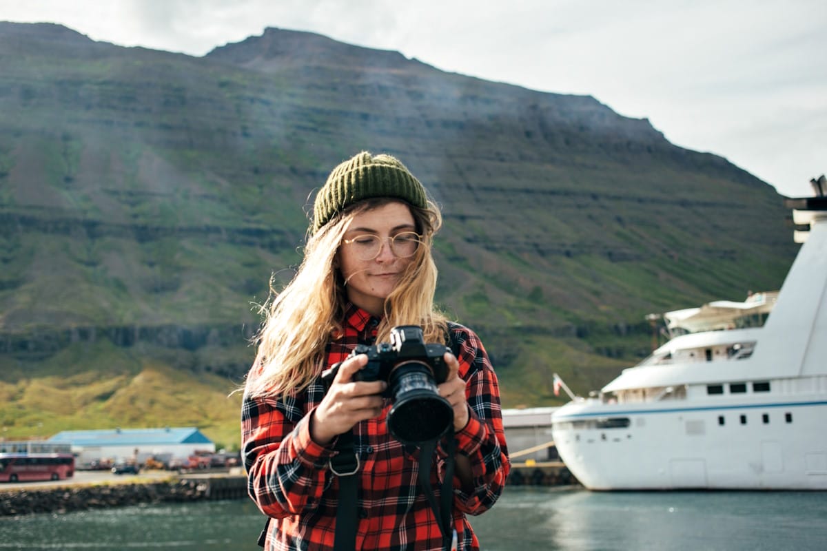 A round woman looking at her camera lens with a mountain and cruise ship in the background